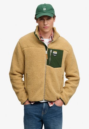 Giovane uomo che indossa una giacca in pile color sabbia con tasca verde sul petto, cappellino da baseball verde, camicia rossa a quadri e jeans blu, in piedi con le mani nelle tasche.