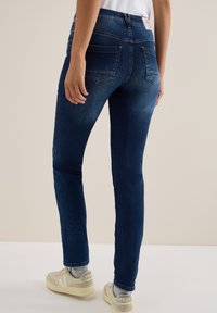 Jeans de mezclilla en azul oscuro, diseño ajustado, cintura a media altura, con bolsillos traseros y una textura ligeramente descolorida. Combinados con zapatillas ligeras.