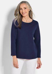 Lächelnde Frau mit langen grauen Haaren, die ein navyblaues langärmliges Oberteil und eine weiße Hose trägt, stehend vor einem schlichten hellen Hintergrund.