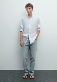 Chemise à rayures blanches, jean bleu clair et tongs noires ; le modèle se tient avec les mains dans les poches devant un fond uni.
