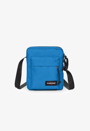 Blue fabric crossbody bag with a zip closure, featuring a front pocket, adjustable black strap, and a branded patch on the front.