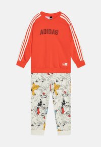 Felpa rossa con logo Adidas bianco e tre strisce bianche sulle maniche, abbinata a pantaloni beige con disegni colorati di Topolino.