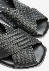 Sandalias de cuero trenzado negro con correas entrecruzadas, punta cuadrada y una suela lisa y plana. Textura notable por el diseño trenzado.