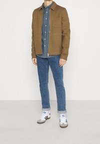 Hnědá bunda na zip s límcem, doplněná modrou košilí na knoflíky, modrými džínami a bílými teniskami s černými doplňky.