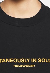 Czarny t-shirt z bawełny z okrągłym dekoltem i wyraźnym żółtym napisem "SPONTANEOUSLY IN SOLID" z "HOLZWEILER" poniżej.