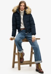 Chaqueta de plumas azul marina con capucha forrada de piel, de corte oversize, combinada con un suéter crema, jeans de mezclilla azul claro y botas marrones con cordones amarillos.