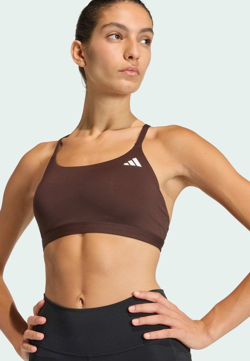 Femme en forme portant un brassière de sport adidas marron et des leggings noirs, posant les mains sur les hanches, regardant sur le côté sur un fond clair.