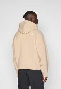 Maglione beige con cappuccio, texture a coste, maniche ampie e un piccolo logo ricamato sulla schiena. Il design presenta una vestibilità rilassata.