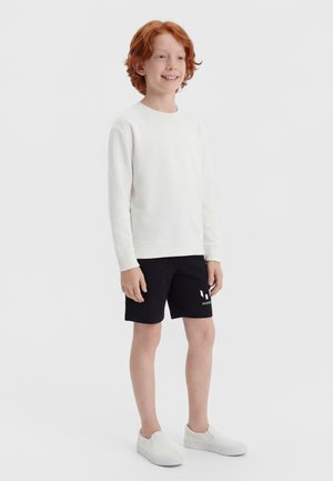 Lächelnder rothaariger Junge steht, trägt ein weißes Langarmshirt, schwarze Shorts mit einem Logo und weiße Slipper auf weißem Hintergrund.