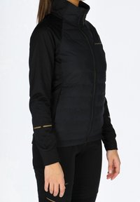 Veste de sport noire avec fermeture éclair, devant matelassé et côtés en tissu contrastant. Comprend des accents dorés sur les manches et le logo.