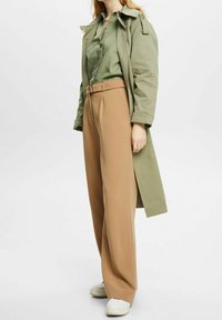 Trench coat verde sopra una camicia verde chiaro, abbinato a pantaloni beije a vita alta e sneakers bianche. Tessuti morbidi e vestibilità rilassata.