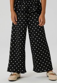 Crne široke hlače s bijelim točkicama, izrađene od teksturiranog materijala s poderanim rubovima. Uparene sa svijetlim ravnim cipelama.