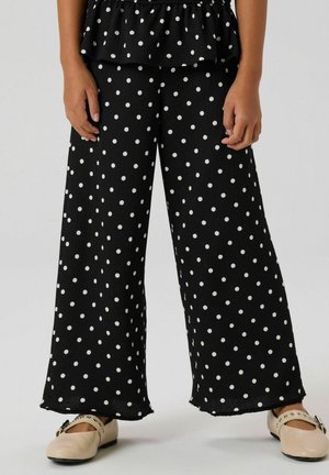 Czarne spodnie o szerokich nogawkach w białe grochy, wykonane z fakturowanego materiału z postrzępionymi lamówkami. W połączeniu z jasnymi, płaskimi butami.