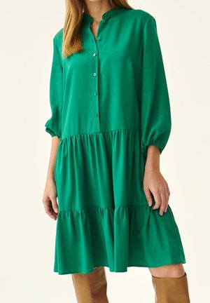 Femme portant une robe verte à manches longues à volants avec des boutons sur le devant, associée à des bottes marron montant jusqu'aux genoux.