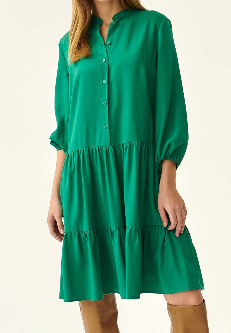 Femme portant une robe verte à manches longues à volants avec des boutons sur le devant, associée à des bottes marron montant jusqu'aux genoux.