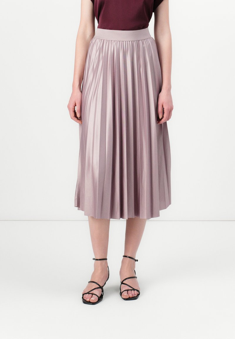 Pleated midi-hame pehmeässä mauve-satinkankaassa, jossa on leveä vyötärönauha, yhdistettynä mustiin hihna-sandaaleihin, yksinkertaisella taustalla.