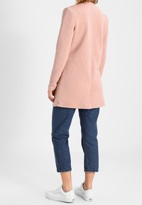 Ljusrosa texturerad kappa med långa ärmar, knälång design, bärs över uppvikta blå jeans och vita sneakers. Anmärkningsvärda sömdetaljer.