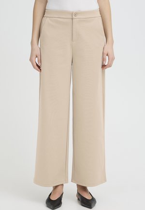 Person trägt beigeweite Hosen, weißes Oberteil und schwarze spitze Ballerinas, steht mit entspannten Händen an den Seiten.