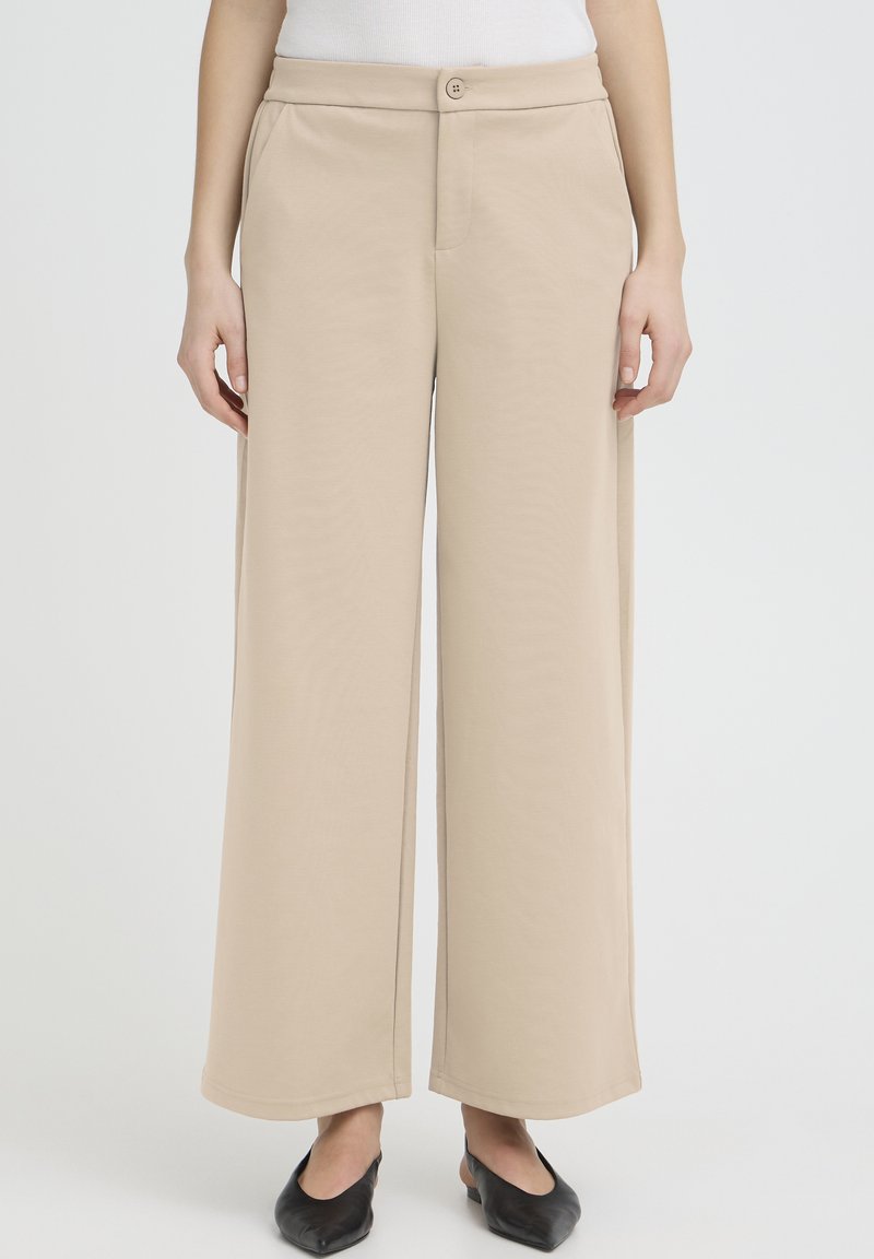 Osoba oblečená v béžových širokých nohaviciach, bielom topánku a čiernych špicatých balerínach, stojaca s uvoľnenými rukami pozdĺž tela.