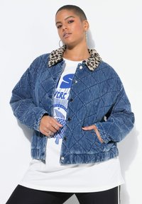 Pikowana kurtka dżinsowa z kołnierzem w panterkę, zapinana na zatrzaski, z dwiema przednimi kieszeniami, noszona na białej koszulce z nadrukiem.