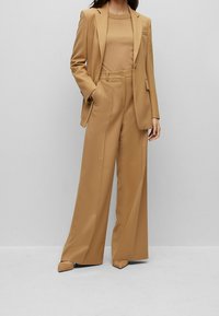 Beżowy garnitur z dopasowaną marynarką i szerokimi spodniami. Gładka faktura, zapięcie na jeden guzik oraz minimalistyczne kieszenie.