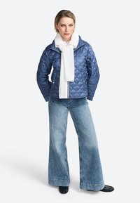 Modra podložena jakna z visokim ovratnikom, v kombinaciji z belim šalom, širokimi denim kavbojkami in črnimi čevlji, na svetli podlagi.