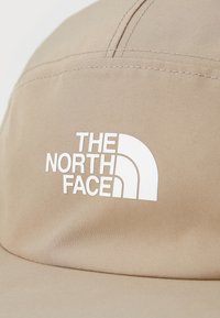 Beige színű, sima textúrájú anyagból készült sapka, amelyen a "THE NORTH FACE" felirat fehér színben és egy stilizált félkupola grafika található.