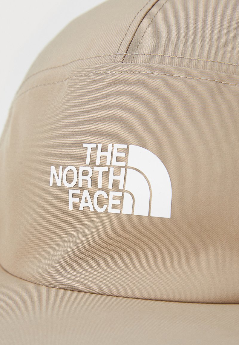 Beige pet gemaakt van stof met een soepele textuur, met een wit logo tekst "THE NORTH FACE" en een gestileerde halve koepel grafiek.