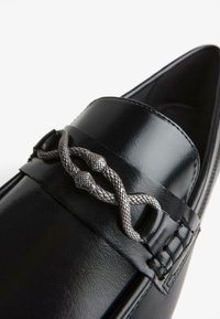 A fekete bőr loafer egy fonott ezüstszínű díszítőelemmel büszkélkedhet a felső részén, sima vonalakkal és hegyes orrformával.