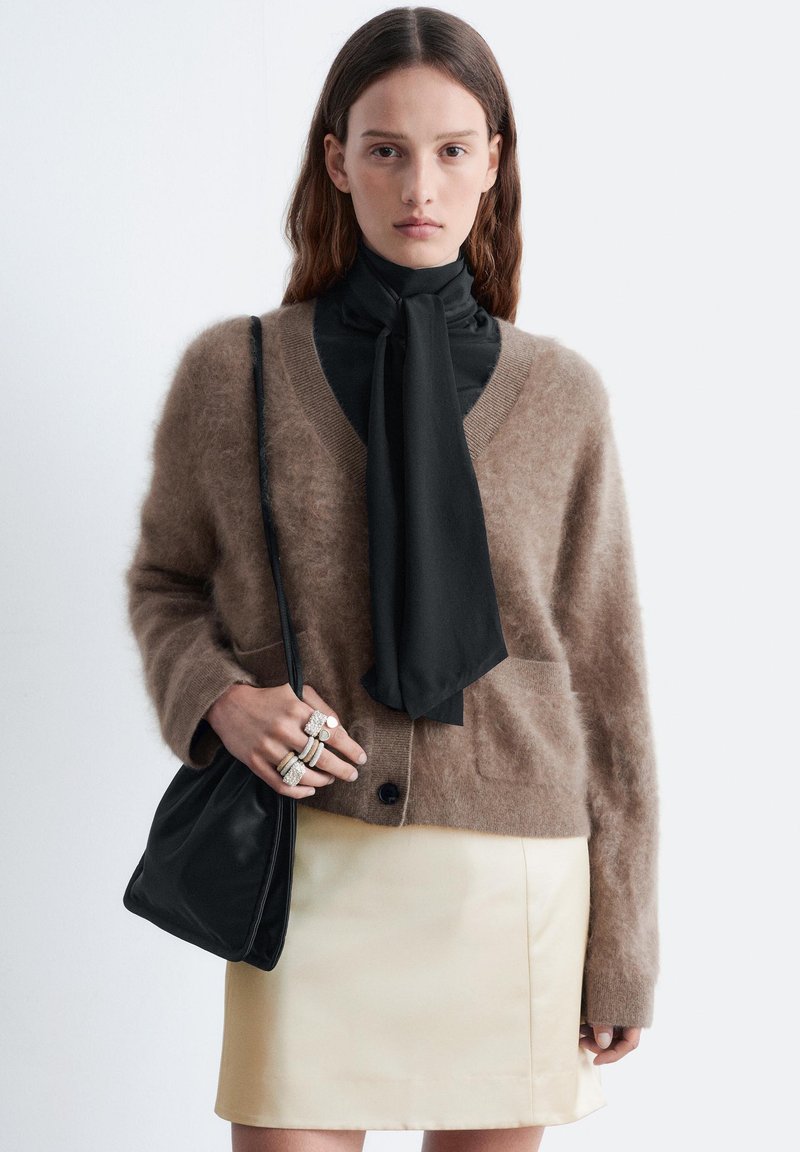 Brauner, flauschiger Cardigan mit Taschen, darüber eine schwarze Bluse mit Krawatte, kombiniert mit einem cremefarbenen Lederrrock und einer schwarzen Tasche in der Hand.