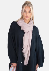 Écharpe texturée rose clair à bords frangés, enroulée de manière décontractée autour du cou, portée par-dessus une chemise noire à boutons ample.