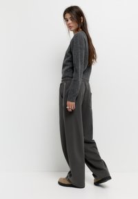 Pulover din lână gri, cu mâneci lungi, asociat cu pantaloni largi de culoare gri închis, cu un tiv elastic și buzunare. Încălțăminte plată de culoare maronie.
