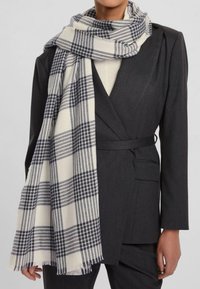 Sciarpa a scacchi in crema e blu marino, realizzata in un tessuto morbido, appoggiata su un blazer scuro su misura con un design strutturato e dettagli con cintura.