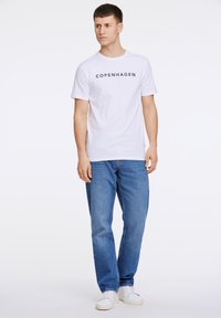 Weißes Baumwoll-T-Shirt mit „KOPENHAGEN“ in Marineblau bedruckt. Kombiniert mit hellblauen Jeans und weißen Sneakers. Schlichtes, lässiges Design.