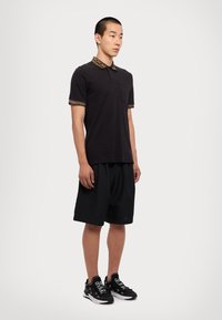 Čierne polo tričko so vzorovaným golierom a rukávmi, v kombinácii s voľnými čiernymi šortkami a čiernymi teniskami. Minimalistický dizajn a čisté línie.