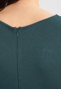 Haut teal à manches courtes avec un décolleté en V et une fermeture éclair au dos, fabriqué à partir d'un tissu doux et lisse avec une finition légèrement texturée.