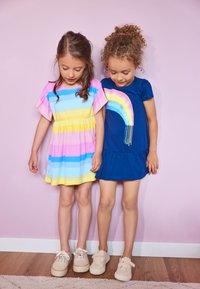 Two children stand side by side. One wears a pink and blue striped dress; the other, a blue dress with a rainbow design. Both wear casual shoes.