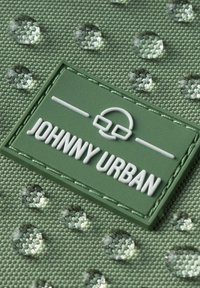 Tessuto verde testurizzato con gocce d'acqua trasparenti, caratterizzato da un'etichetta in gomma rettangolare impressa con "JOHNNY URBAN" e un elemento grafico.