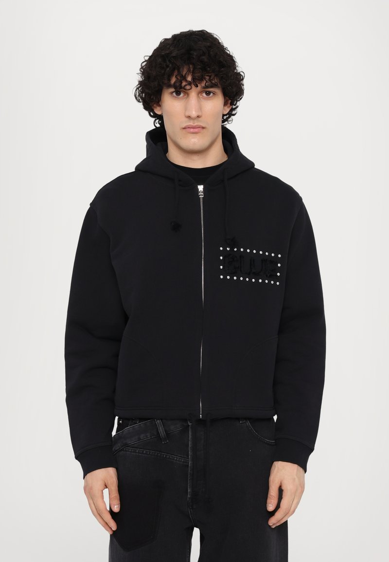 Veste à capuche zippée noire avec une poche avant, présentant un design texturé "BLUE" rehaussé de clous argentés sur la zone gauche de la poitrine.