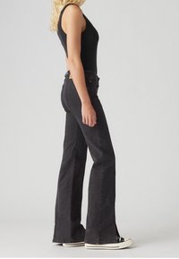 Pantalones de mezclilla negros acampanados con cintura alta, cuentan con una abertura lateral en el dobladillo y costuras en contraste. Combinados con una blusa negra y zapatillas.