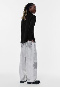 Schwarzes Langarmshirt, kombiniert mit lockeren, hellgrauen Denim-Jeans mit einem ausgewaschenen Look und großen Taschen. Schwarze Schuhe runden das Outfit ab.
