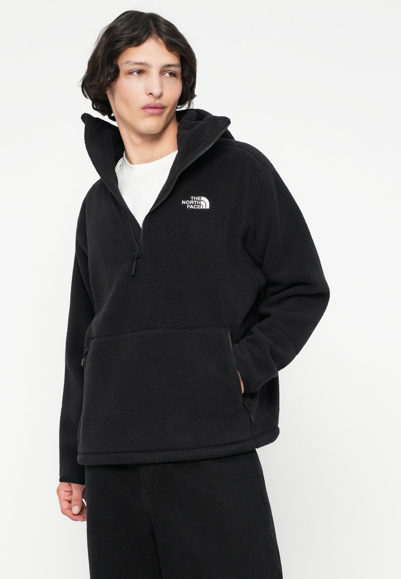 Junger Mann, der einen schwarzen Fleece-Pullover mit einem Viertelreißverschluss und dem Logo von The North Face trägt, seitlich schaut und eine Hand in der Tasche hat.