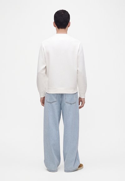Homme portant une chemise blanche à manches longues et un jean large bleu clair, debout de dos devant un fond uni.