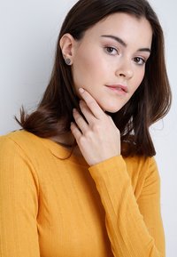 Hranaté, šedé náušnice na modelke s stredne hnedými vlasmi. Má na sebe textúrovaný, dlhý rukáv, horčicovožltý top a za ňou je neutrálny pozadie.