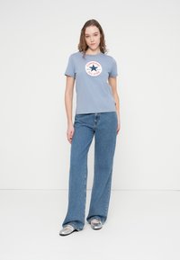 Ανοιχτό γαλάζιο βαμβακερό t-shirt με στρογγυλή λαιμόκοψη και γραφικό λογότυπο, συνδυασμένο με τζιν παντελόνι υψηλής μέσης και φαρδιάς γραμμής σε μπλε χρώμα και ασημί παπούτσια τύπου slip-on.