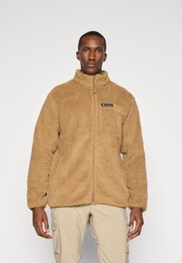 Tanskleurige fleecjas met een hoge kraag, ritsluiting en een linkerkant borstzak met een logopatch. Gladde textuur en een ontspannen pasvorm.