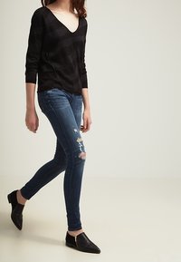 Svart V-ringad tröja med horisontella ränder, kombinerad med blå slitna skinny jeans och svarta platta skor med sidoflodrande detalj.