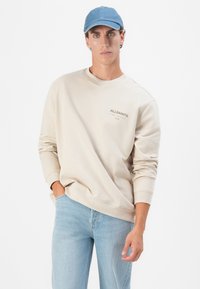 Sweat-shirt beige avec un col rond et des manches à revers, arborant un petit logo. Assorti à un jean bleu clair et une casquette bleu clair.