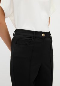 Zwarte broek met een soepele textuur, een slim fit, zijzakken en een metallic gouden knoop in de taille.
