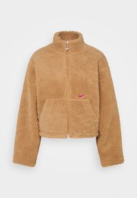 Beige imitationpäls zip-up jacka med hög krage, framfickor och en rosa Nike-logotyp på vänster bröst, med en strukturerad yta.